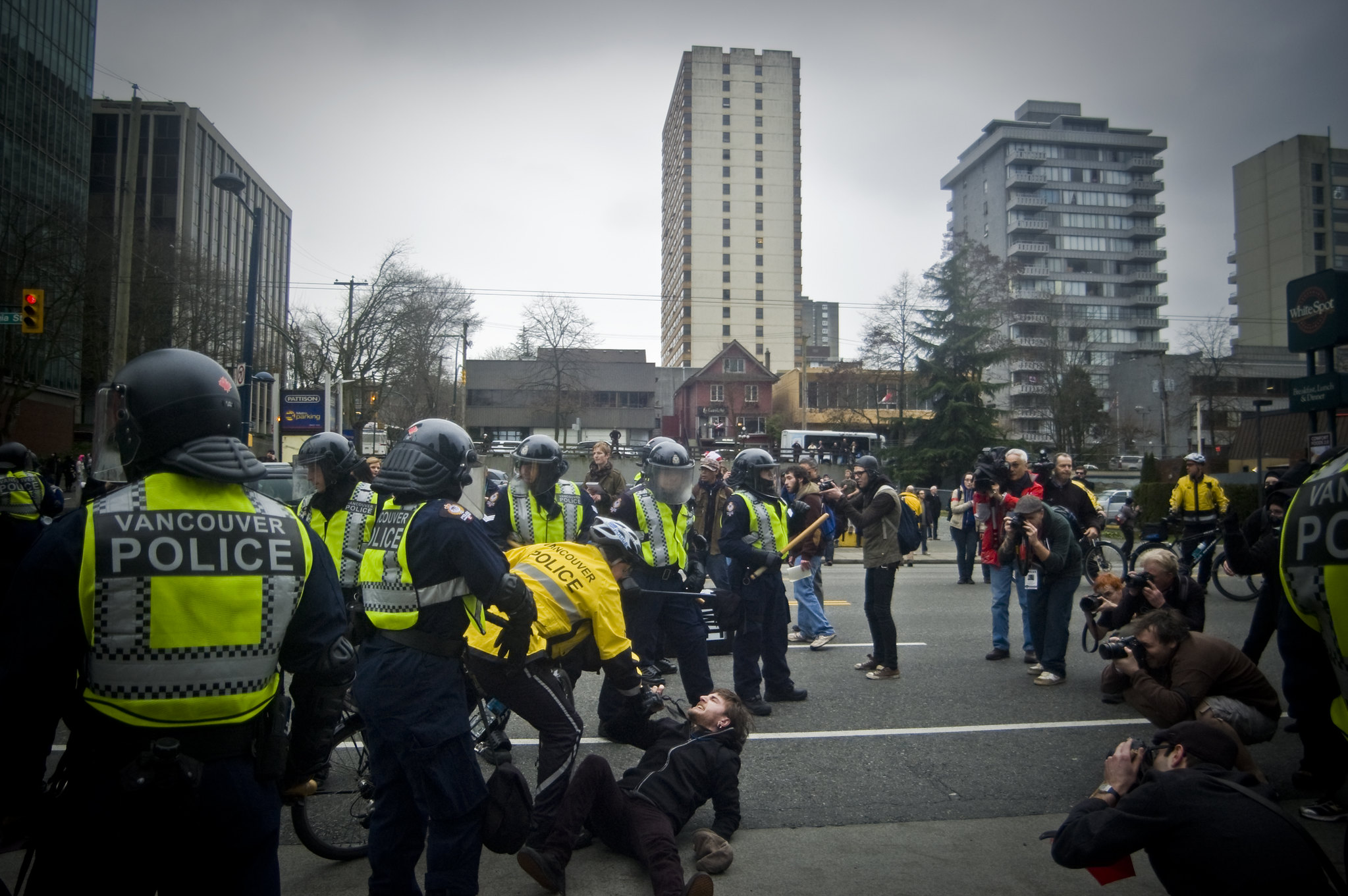 A police officer violently handling an unmasked black bloc person on the ground, with a ring of photographers surrounding