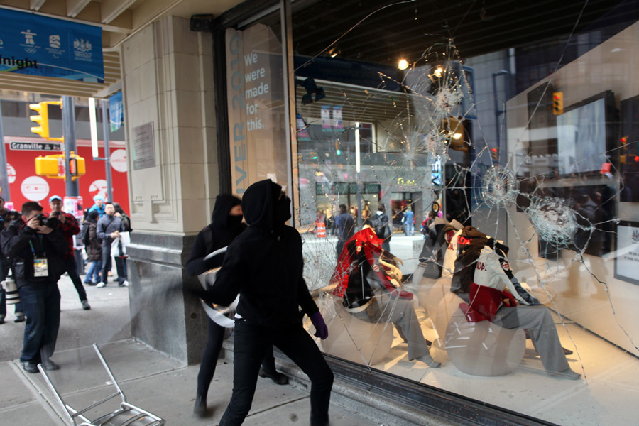 Two black bloc demonstrators in front of an HBC display window, they are engaged in breaking it, spiderweb cracks are visible